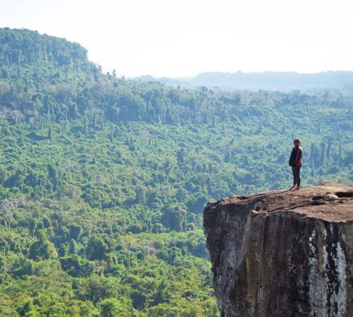 A tourist standing on a rocky cliff viewpoint overlooking the vast green jungle at Phnom Kulen National Park near Blanc Smith Hotel in Siem Reap.