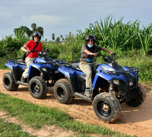 Two tourists wearing safety helmets while riding blue quad bikes on a dirt path near Blanc Smith Hotel in Siem Reap.