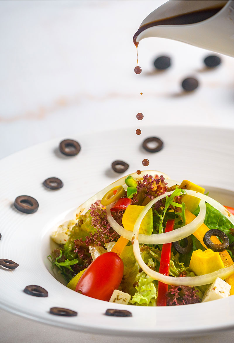 Fresh Mediterranean Greek salad with olives, feta cheese, and balsamic dressing being poured at Blanc Smith Hotel restaurant in Siem Reap.